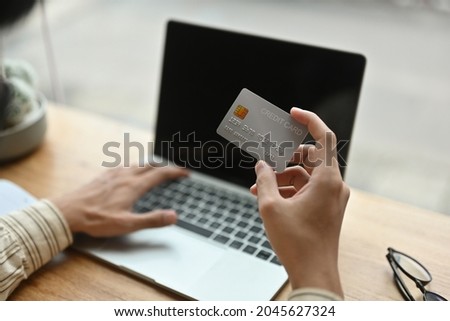 Cropped image of hands holding a credit card in front of a laptop computer at the wooden counter. Paying by credit card concept.