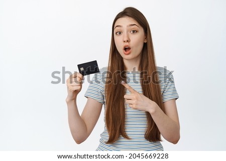 Curious girl points at credit card, asking question about discount or bank, stands against white background