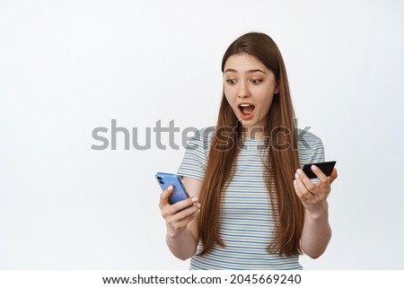 Girl looks surprised at her mobile phone, holds credit card. Concept of online shopping on smartphone, white background