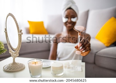 Beautiful woman using stone facial roller. Photo of african american woman after shower on home background. Beauty and skin care concept. Woman doing self massage with rose quartz face roller
