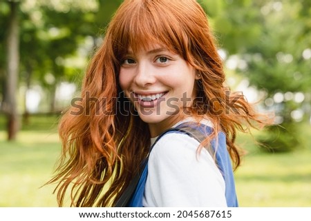 Close up portrait of a young caucasian woman girl teenager student schoolgirl with ginger red hair and toothy smile walking in park forest outdoors looking at camera. Royalty-Free Stock Photo #2045687615