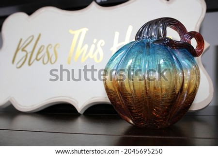 A close up, isolated, Fall background image. This Autumn scene is of a teal blue and gold glass pumpkin that is sitting in front of a sign that reads "Bless this house" in gold lettering.