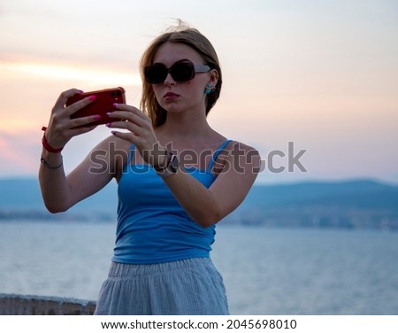 A blonde girl in dark glasses takes pictures of herself on her phone against the background of the sea and the sunset.