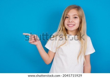 beautiful caucasian little girl wearing white T-shirt over blue background points at copy space and advertises something, advices best price.