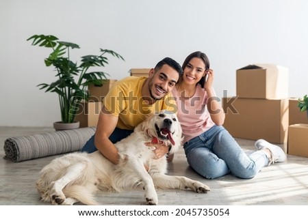 Full length portrait of happy young diverse couple with their dog posing on floor of new home on moving day. Millennial homeowners with cute pet sitting among cardboard boxes, relocating to new house Royalty-Free Stock Photo #2045735054