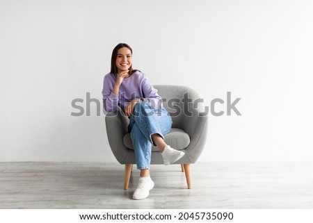 Full length portrait of happy young woman in casual wear smiling at camera, sitting in comfy armchair against white studio wall. Lovely millennial lady posing, relaxing indoors Royalty-Free Stock Photo #2045735090