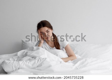 Sad european millennial female, worried, suffers from insomnia and headache. Lady sits on bed in bedroom at home, copy space. Migraines, health and mental problems, lack of sleep and depression Royalty-Free Stock Photo #2045735228