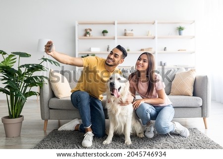 Happy interracial couple taking selfie with their dog while sitting on soft carpet at home, full length. Positive Arab guy and his girlfriend making photo with cute pet in living room