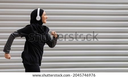 Fit muslim woman in modest sportswear and wireless headphones running outdoors, side view shot in motion of athletic islamic female runner in hijab doing sports outside, panorama with copy space Royalty-Free Stock Photo #2045746976