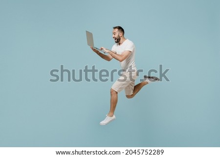 Full length side view young overjoyed man 20s in casual white t-shirt jump high run fast hurrying up hold closed laptop pc computer isolated on plain pastel light blue color background studio portrait Royalty-Free Stock Photo #2045752289