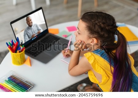 Preschooler girl looks at computer screen and studies with young black male teacher at home interior. Modern online education, tutoring and lesson during covid-19 quarantine. Knowledge and learning