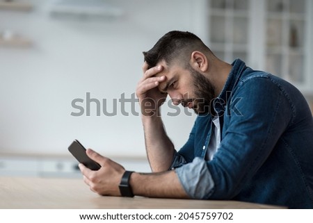 Waiting for call, problems and difficulties, stress and no answer. Unhappy sad disappointed young bearded guy looks at smartphone and presses hand to head, sits at table in kitchen interior, profile Royalty-Free Stock Photo #2045759702