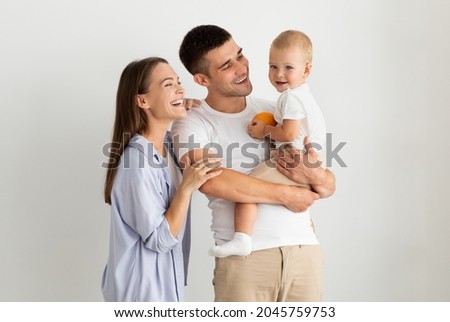 Happy Family Of Three With Cute Infant Baby Posing Over White Background, Cheerful Young Parents And Their Adorable Little Child Embracing And Laughing, Enjoying Spending Time Together, Copy Space