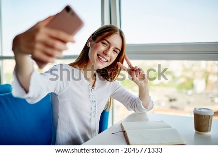 Joyful female influencer in bluetooth earbuds showing v sign while shooting video vlog via app on mobile technology, portrait of happy Caucasian hipster girl in headphones creating selfie content