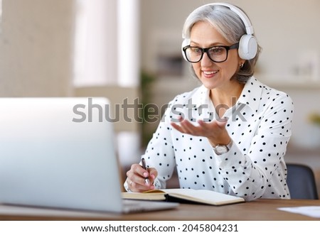 Beautiful smiling mature woman in headphones during online meeting on laptop with colleagues at home, senior female participating in virtual class, learning foreign language remotely at home Royalty-Free Stock Photo #2045804231