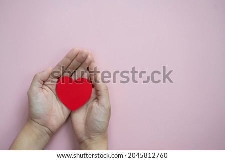 Hands holding red heart on pastel pink background.