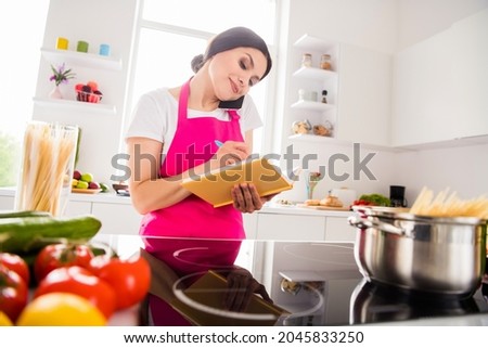 Photo portrait woman in pink apron writing recipe in blocknote cooking pasta talking on cellphone