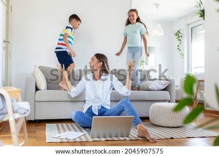 Tired young mother sitting on the floor and working with laptop and documents while little kids and jumping on the sofa and having fun and making noise