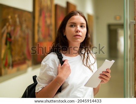 Portrait of a focused girl standing in the museum hall next to an exhibit located in a glass cabinet, with a booklet of the ..exhibition program Royalty-Free Stock Photo #2045914433