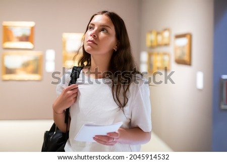Focused young girl visitor holds a booklet with an exhibition program while admiring the paintings in the museum Royalty-Free Stock Photo #2045914532