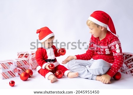 a little girl in a Santa costume and a red cap looks at gifts with a boy in a red sweater on a white background, Christmas, new year