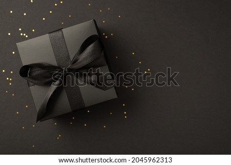 Top view photo of black giftbox with black ribbon bow and golden confetti on isolated black background with copyspace
