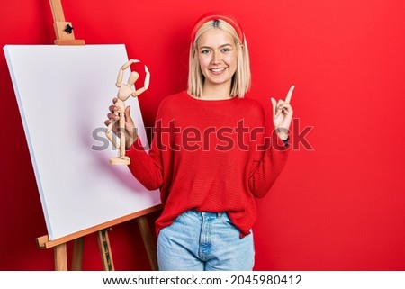 Beautiful blonde woman holding small wooden manikin smiling happy pointing with hand and finger to the side 