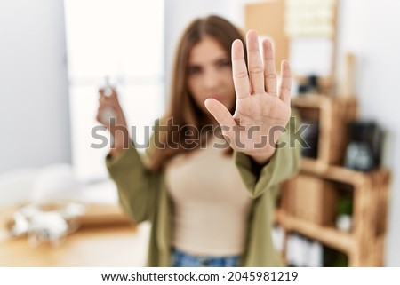 Young brunette woman working at architecture office holding house keys with open hand doing stop sign with serious and confident expression, defense gesture 