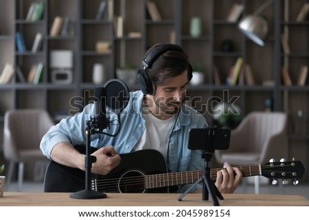 Man in headphones playing acoustic guitar, recording video on smartphone standing on tripod, using professional microphone, blogger or music teacher shooting course in home studio, sitting at desk