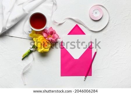 Cup of tea, beautiful flowers and blank card with envelope on light background