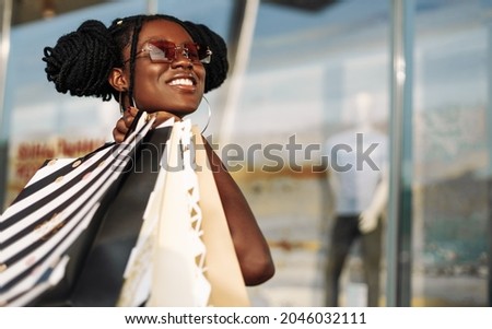Young smiling african american woman, wearing sunglasses, with shopping bags in mall, shopping, black friday, discounts, sale concept Royalty-Free Stock Photo #2046032111