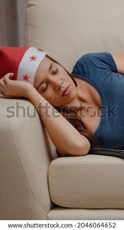 Close up of woman sleeping on couch at television while having santa hat on head for christmas eve celebration. Adult laying on sofa falling asleep with bowl of chips on holiday