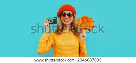 Autumn portrait of happy smiling young woman with film camera and yellow maple leaves wearing a sweater, french beret on blue background