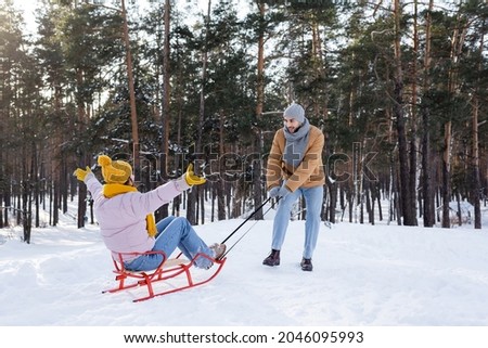 Smiling man pulling girlfriend on sled on snow in park