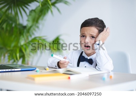 Elementary school child in a bad mood writes in a notebook. White desk, laptop. Large houseplant, palm tree on the background. Daylight. Concept for study, education, unhappy student, homework problem