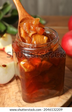 Spoon with tasty apple jam in glass jar on wooden board, closeup