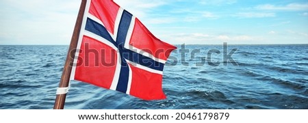 Flag of Norway against cloudy blue sky, close-up. North sea in the background. Travel destinations, national identity, symbolism, patriotism
