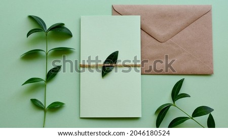 Mockup of craft paper envelope with blank green card on light green background with plants leaves close-up. Wedding, Happy Birthday, Valentine's day, Mother's Day greeting, invitation card concept.