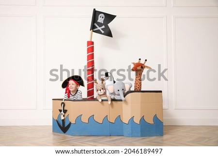 Cute little boy playing with toys in pirate cardboard ship near white wall indoors Royalty-Free Stock Photo #2046189497