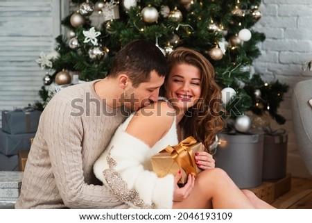 A happy couple in love celebrates a new year or Christmas. The couple sits on the floor near the Christmas tree in a home atmosphere and exchange gifts.