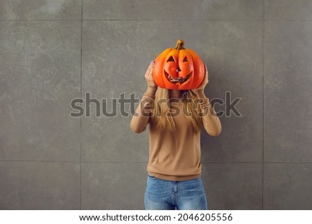 Woman holding orange pumpkin she has made for Halloween party. Young lady hiding face behind carved pumpkin. Studio shot of teenage girl with Jack-o-lantern instead of head standing against grey wall