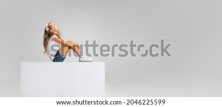 Dreaming, looking up. Portrait of little cute girl in casual clothes sitting on big box isolated on white studio background. Happy, joyful childhood, kids fashion, emotions, facial expression concept