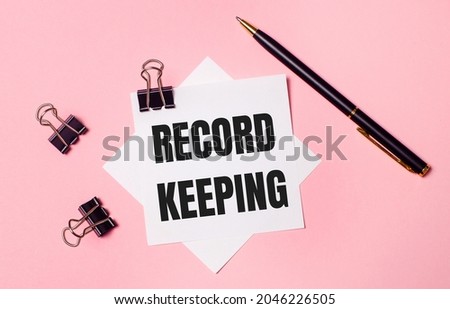 On a light pink background, black paper clips, black pen and white note paper with the words RECORD KEEPING. Flat lay
