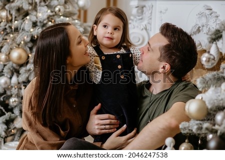 Happy dad, mom and daughter are enjoying the New Year and Christmas holidays. Dad, mom and daughter are sitting against the background of a stylishly decorated Christmas tree and enjoy the holiday