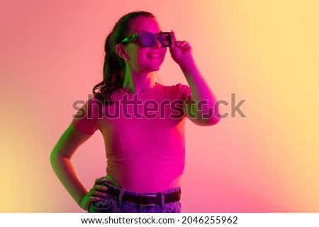 One young caucasian girl in sunglases smiling and posing isolated over gradient studio background in neon lights. Youth culture. Concept of human emotions, facial expression, beauty. Copy space for ad