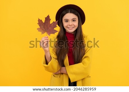 smiling teen girl in hat and coat with autumn maple leaf on yellow background, beauty
