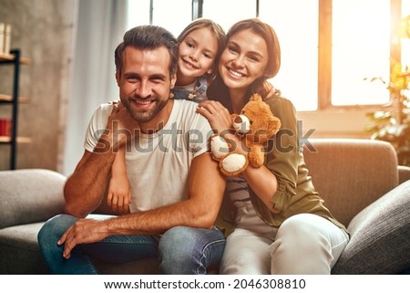 Happy dad and mom with their cute daughter and teddy bear hug and have fun sitting on the sofa in the living room at home. Royalty-Free Stock Photo #2046308810