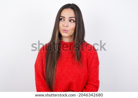young beautiful brunette wearing red knitted sweater over a white wall, looks pensively aside, plans actions after university, imagines what to do Thinks over about new project.