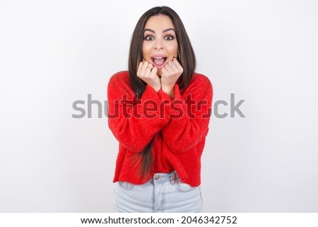 Indoor closeup of young beautiful brunette wearing red knitted sweater over a white wall practicing yoga and meditation, holding palms together in namaste, looking calm, relaxed and peaceful.