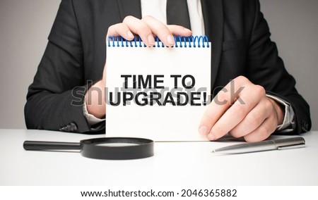 man writes in a notebook with a silver pen and hand holding card with text Time to upgrade. grey background, front view. business and education concept.